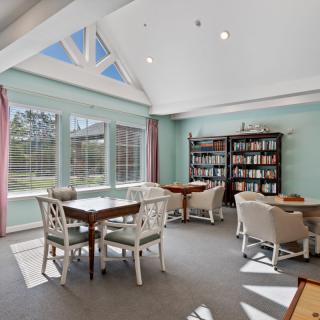 A cozy reading room featuring bookshelves, tables, and chairs with abundant natural light from large windows.
