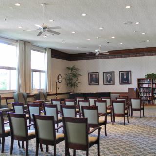 An empty conference room with arranged chairs and bookshelves, illuminated by natural light from large windows.