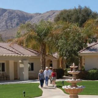 A group of people walking along a pathway near a fountain with houses and mountains in the background.