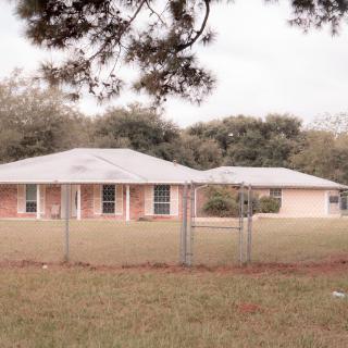 A single-story suburban house with a fenced yard and surrounding trees.
