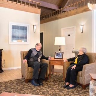 An elderly man and woman sit in a cozy living room, engaged in conversation.
