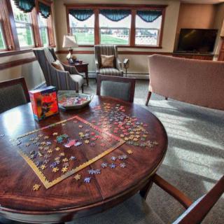 A cozy living room with a puzzle in progress on a round wooden table surrounded by comfortable seating and large windows.