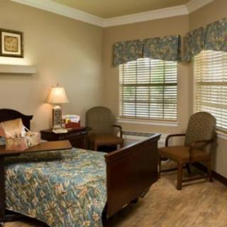 A cozy bedroom with a neatly made bed, two chairs, a lamp on a nightstand, and windows with floral curtains.