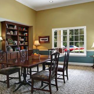 A cozy dining room featuring a wooden table and chairs, with a bookshelf and large window.