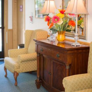 A cozy living room corner with upholstered chairs, a wooden cabinet topped with vibrant flowers and lamps.