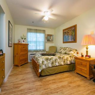 A cozy bedroom with wooden flooring, featuring a bed, dresser, nightstand with a lamp, and wall decor.
