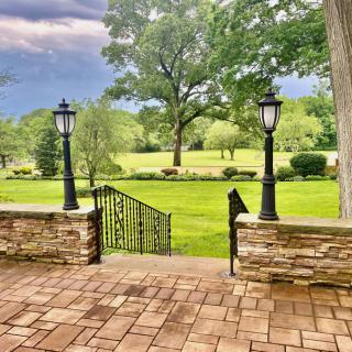 A scenic view from a stone patio overlooking a lush green lawn with trees and lantern lighting.