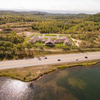 Aerial view of a building complex surrounded by lush greenery, with a road and vehicles in front, adjacent to a body of water.
