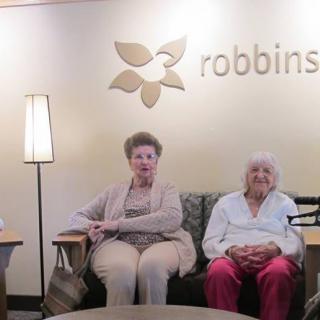 Two elderly individuals seated in a retirement home lounge area under a logo.