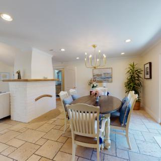 A cozy dining room with a wooden table and chairs, a chandelier overhead, tiled flooring, and decorative plants and artwork.