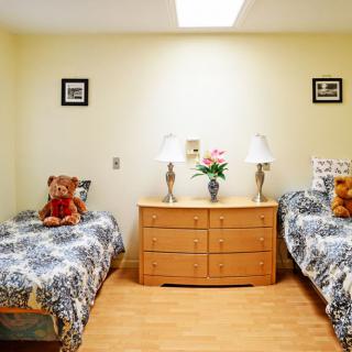 A cozy bedroom featuring twin beds with matching bedding, decorated with teddy bears and a wooden dresser centered between them, adorned with lamps and flowers.