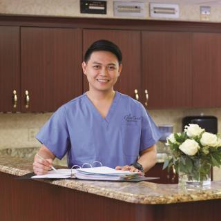 A medical professional in blue scrubs stands at a reception desk with a clipboard.
