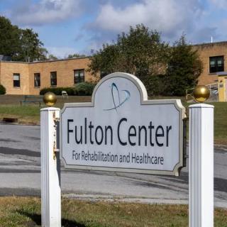 A sign for the Fulton Center for Rehabilitation and Healthcare stands in front of a brick building under a partly cloudy sky.