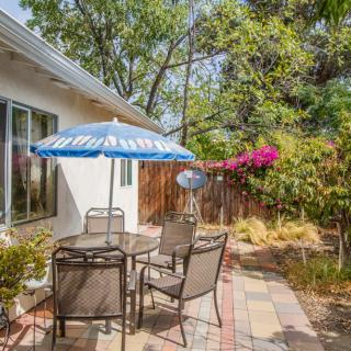A cozy outdoor patio area with a table, chairs, and an umbrella, surrounded by greenery and flowers.