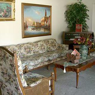 A vintage living room featuring floral-patterned furniture, artwork on the wall, a piano, and decorative plants.