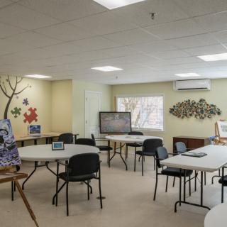 A community meeting room with tables and chairs arranged for group activities, featuring wall decorations and natural light from a window.