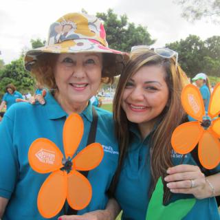 Two women smiling and holding orange flower decorations at an outdoor event.