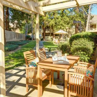 A sunny backyard with wooden patio furniture under a pergola, surrounded by lush greenery.