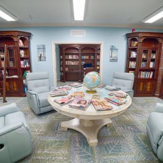 A cozy library corner with reclining chairs, a central table displaying magazines, and wooden bookshelves lining the walls.