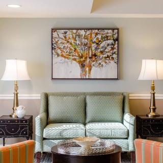 A cozy living room with a green sofa, framed artwork of a tree, and elegant lamps on side tables.