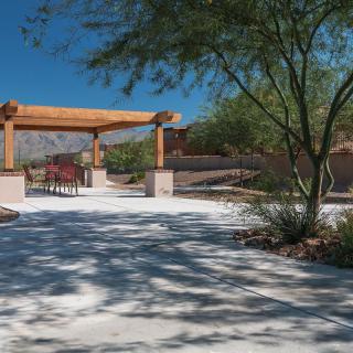A serene park with a pathway leading under a wooden pergola, surrounded by trees and desert landscaping.