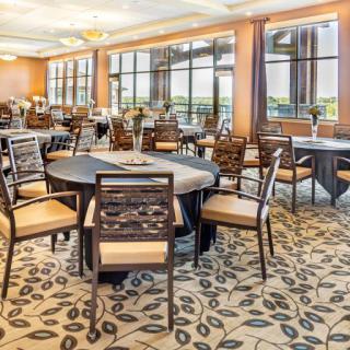 A spacious dining area with round tables and cushioned chairs, featuring large windows and a patterned carpet.