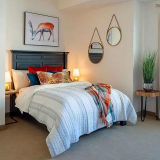A stylish bedroom featuring a neatly made bed with striped linens, decorative pillows, and artwork above the headboard. Two round mirrors are mounted on the wall, and a potted plant sits on a side table.