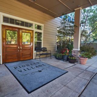 Entrance of a building with wooden doors, a mat labeled 'Pioneer Village,' and potted plants.