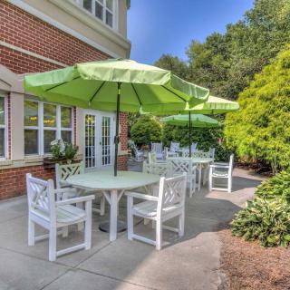 A sunny patio area with green umbrellas shading tables and white chairs beside a brick building, surrounded by lush greenery.