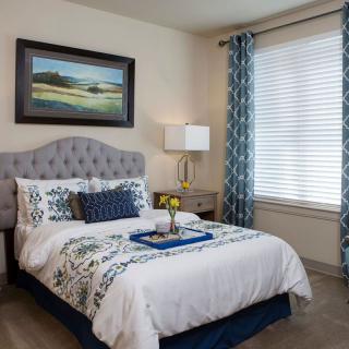 A cozy and stylishly decorated bedroom featuring a neatly made bed with patterned bedding, a nightstand with a lamp, and a large window with blue curtains.