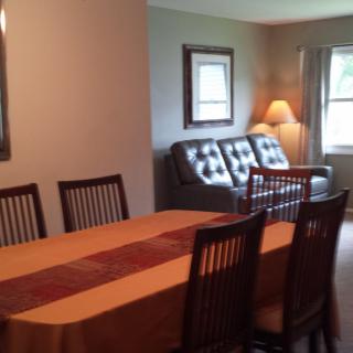 A cozy living and dining area featuring a long table with chairs, a sofa, a lamp, and windows providing natural light.