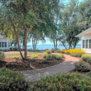 A serene garden pathway leads to a picturesque lakeside view, surrounded by lush greenery and charming buildings.