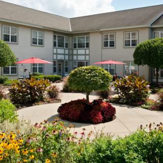 A beautifully landscaped courtyard with colorful flowers, trimmed trees, and sun umbrellas in an outdoor seating area next to a building.