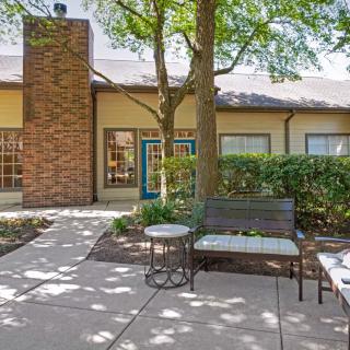A cozy patio area with a bench and table, surrounded by greenery and trees in front of a house with a brick chimney.