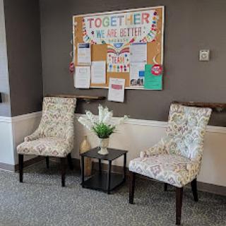 A cozy waiting area featuring patterned chairs, a small table with floral decor, and a colorful bulletin board.