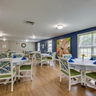 A bright and neatly arranged dining room with multiple tables and chairs, featuring decorative wall art and large windows.