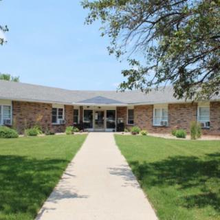 A brick residential building with a pathway leading to the entrance, surrounded by a green lawn and trees.
