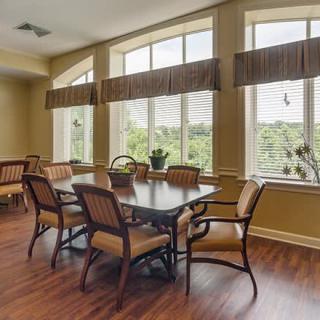 A well-lit dining room with large windows and a wooden table surrounded by chairs.