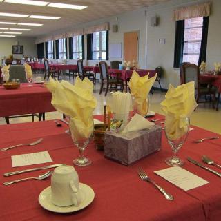 A neatly set dining table in a large dining hall with red tablecloths and yellow folded napkins.