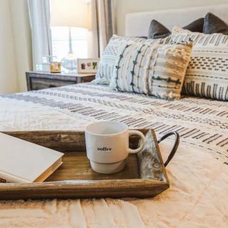 A cozy bedroom setting featuring a neatly arranged bed with decorative pillows and a tray holding a coffee mug and notebook.