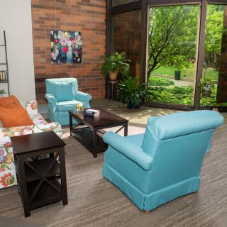 A cozy living room featuring blue armchairs, a floral sofa, and a large window overlooking greenery.