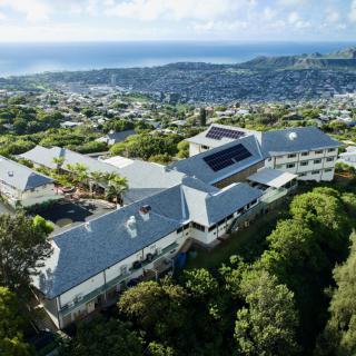 Aerial view of a large building complex surrounded by lush greenery, with a coastal cityscape and ocean in the background.