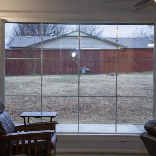 A view of the backyard through a large window from inside a room, featuring chairs and a side table.