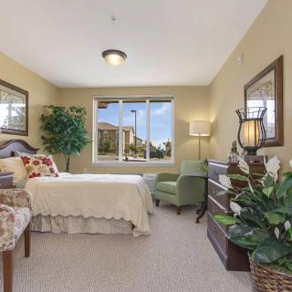 A cozy bedroom with a bed, armchair, dresser, and large window letting in natural light.
