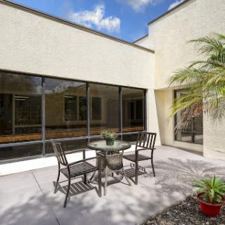 A serene outdoor patio area featuring a glass table and chairs, surrounded by greenery under a blue sky.