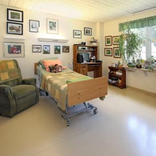 A cozy hospital room with a bed, recliner, and various decorations including framed pictures and plants.