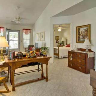 A cozy living room with traditional furniture, featuring a desk and decorations. The room connects to a bedroom, visible through an open doorway.