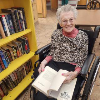 An elderly woman sitting in a wheelchair, enjoying a book in a library setting.