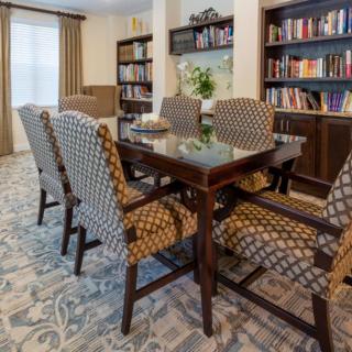 A dining area with a table and patterned chairs, surrounded by bookshelves and a carpeted floor.