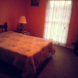 A cozy bedroom with a bed, nightstand, and lamp beside a window with sheer curtains.